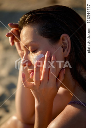 Cream smear. Beauty close up portrait of young woman with healthy skin is applying a skincare product. Girl with suntan spf lotion and cosmetics model face with moisturizing cream, sunscreen on cheek. Cream smear. Beauty close up portrait of young woman with healthy skin is applying a skincare product. Girl with suntan spf lotion and cosmetics model face with moisturizing cream, sunscreen on cheek. 128544717