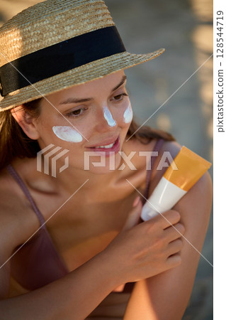 Smiling happy woman applying sunscreen to her face on the beach at sunset. Cream, moisturizer and facial woman in skincare, beauty or cosmetics. Smiling happy woman applying sunscreen to her face on the beach at sunset. Cream, moisturizer and facial woman in skincare, beauty or cosmetics. 128544719