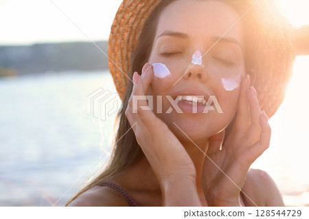 A close-up portrait of a young, happy woman applying sunscreen to her cheeks. Beauty woman with suntan spf and skincare cosmetics model face portrait, woman with moisturising cream, sunscreen product. 128544729