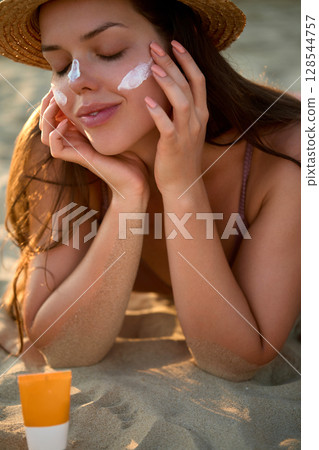 A beautiful woman smiles, applying sunscreen on skin. Skin and Body Care. Sun protection.  A female in a hat smears moisturizing lotion or suntan. 128544757