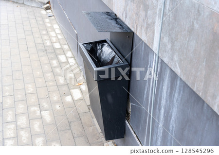 A black trash bin is installed on a wall next to a stone pathway, promoting cleanliness in the urban area A black trash bin is installed on a wall next to a stone pathway, promoting cleanliness in the urban area 128545296