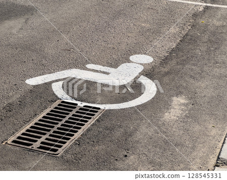 A parking space marked with a disability symbol is positioned directly above a drain in a public lot during bright daylight A parking space marked with a disability symbol is positioned directly above a drain in a public lot during bright daylight 128545331
