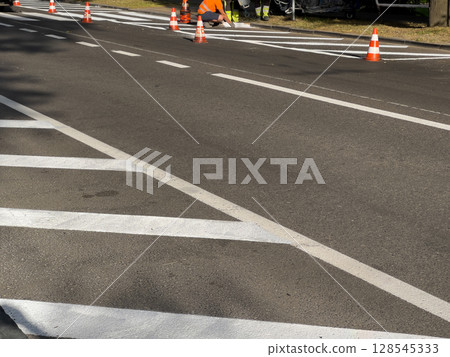 Workers in safety gear apply fresh paint on road lines with traffic cones placed around their work area in the morning light Workers in safety gear apply fresh paint on road lines with traffic cones placed around their work area in the morning light 128545333