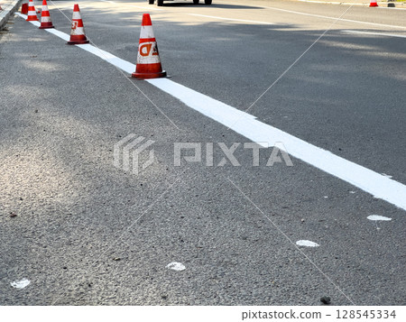 Workers are applying fresh white paint on a city road while traffic cones ensure safety and direct vehicles away from the work area Workers are applying fresh white paint on a city road while traffic cones ensure safety and direct vehicles away from the work area 128545334