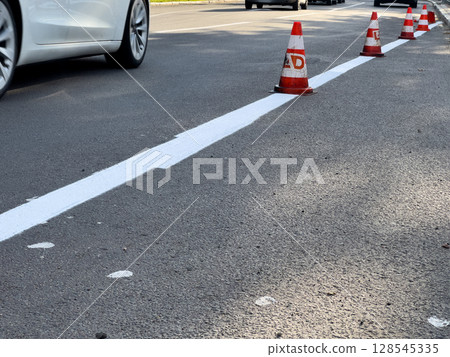 Traffic cones line a freshly painted white line on a busy urban roadway, indicating active road work and maintaining safety for drivers Traffic cones line a freshly painted white line on a busy urban roadway, indicating active road work and maintaining safety for drivers 128545335