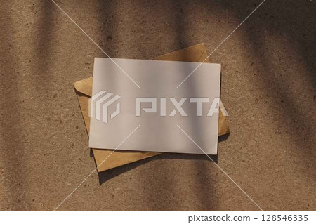 Minimalist horizontal blank greeting card invitation mockup, craft paper envelope. Neutral stone background. Soft natural floral shadows, warm sunlight. Flat lay, top view. Elegant branding stationery 128546335