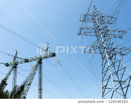 Electric power transmission towers with high-voltage lines against clear blue sky, metal lattice structures for energy distribution, industrial infrastructure in outdoor urban environment 128546383