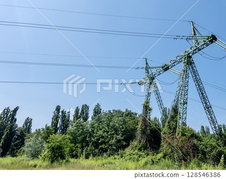 Electric power transmission towers with high-voltage lines against clear blue sky, metal lattice structures for energy distribution, industrial infrastructure in outdoor urban environment 128546386