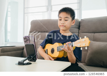 A young boy plays a ukulele on the couch with a smartphone mounted in front for online music tutorials. The home-based learning concept emphasizes modern teaching tools in creative education. A young boy plays a ukulele on the couch with a smartphone mounted in front for online music tutorials. The home-based learning concept emphasizes modern teaching tools in creative education. 128546965