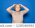Caucasian woman in her 70s wearing a dark beige sweater scratching her head worriedly trying to remember something important, isolated over blue background. 128546992