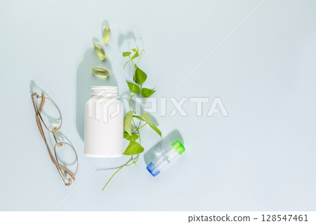 Mockup bottle with natural pills or vitamins for eye health on blue background. White bottle with healthy supplements in gel capsules with glasses and contact lenses. Good vision concept. Copy space 128547461