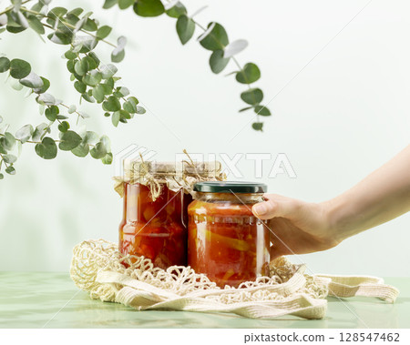 Probiotic foods. Sustainable still life with woman hand hold pickled or fermented vegetables. Lecho and ajvar in glass jars on mesh bag on blue background. Home food preserving or canning Probiotic foods. Sustainable still life with woman hand hold pickled or fermented vegetables. Lecho and ajvar in glass jars on mesh bag on blue background. Home food preserving or canning 128547462