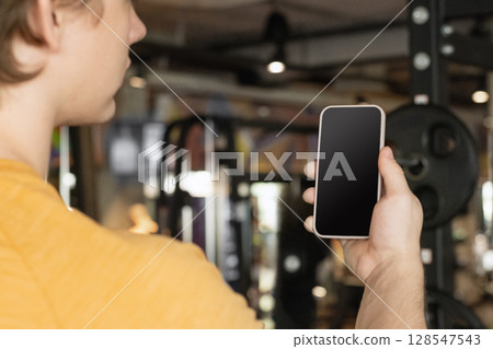 Athlete standing and displaying smartphone with white blank screen in gym setting. Mock up. Space for design. 128547543