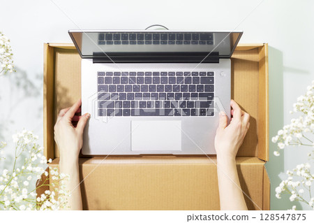 Buying and delivering new laptop. Romantic overhead shot of womans hands holding laptop in cardboard box on blue background with white spring flowers and shadows. Flat lay 128547875