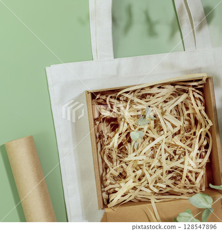 Eco friendly packaging concept. Cardboard box with shredded paper on textile shopping bag on green background with green leaves. Eco packaging goods from online stores and delivery 128547896