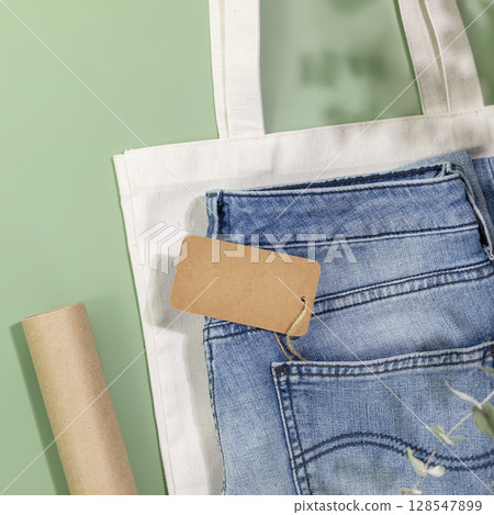 Sustainable flat lay with jeans with empty craft label, textile bag and packing paper on green background. Eco friendly clothing shop. Casual clothes store, shopping, sale and denim shop concept Sustainable flat lay with jeans with empty craft label, textile bag and packing paper on green background. Eco friendly clothing shop. Casual clothes store, shopping, sale and denim shop concept 128547899