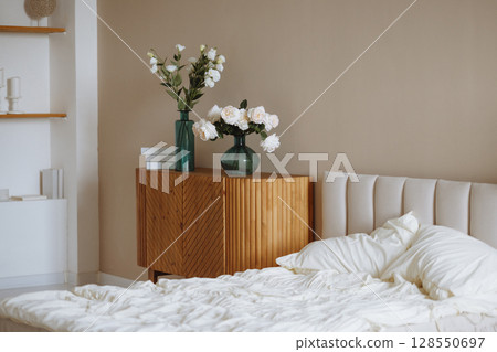 Bright bedroom with unmade white bed, wooden cabinet and flowers in glass vases on beige wall Bright bedroom with unmade white bed, wooden cabinet and flowers in glass vases on beige wall 128550697
