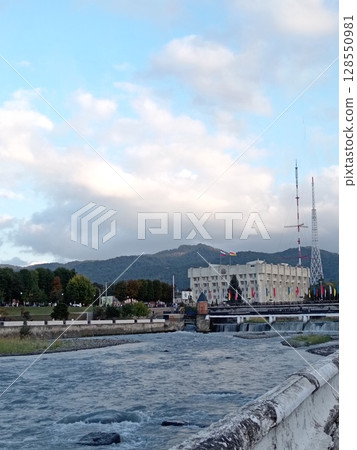 Scenic urban landscape along Terek River: Vladikavkaz City Hall standing proudly on the embankment, Caucasus mountains in background 128550981