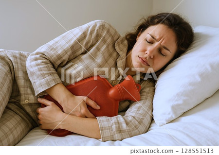 Young woman in pain lying in bed with hot water bottle pressed to stomach, suffering from abdominal cramps or menstrual pain. Concept of home remedy, stomach ache relief, sick day 128551513