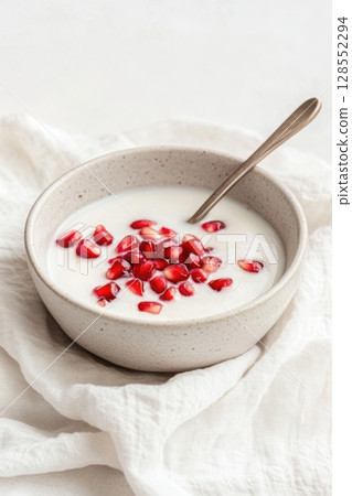 Soft neutral food composition with coconut milk and gently desaturated pomegranate seeds in a minimalist bowl on a warm white backdrop. 128552294