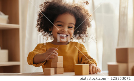 Smiling Black toddler girl plays with wooden blocks at a low table in a cozy room filled with sunlight and natural textures. 128553071