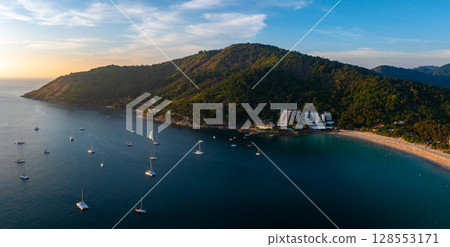 Aerial view of Naiharn Beach in Phuket, Thailand, featuring a crescent shaped shoreline, lush green hills, anchored boats, and warm evening light. Aerial view of Naiharn Beach in Phuket, Thailand, featuring a crescent shaped shoreline, lush green hills, anchored boats, and warm evening light. 128553171