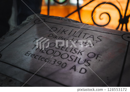Close up of a marble plaque inscribed with details of the Obelisk of Theodosius, framed by ornate wrought iron railing and warm orange lighting. 128553172