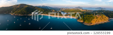 Aerial view of Naiharn Beach in Phuket, Thailand, showing turquoise waters with sailboats, lush green hills, and a hilltop structure in the distance. 128553199