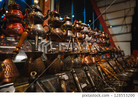 Copper and metal coffee pots with intricate engravings displayed on a glass counter, surrounded by loose tea and spices in a market in Istanbul, Turkey. 128553200