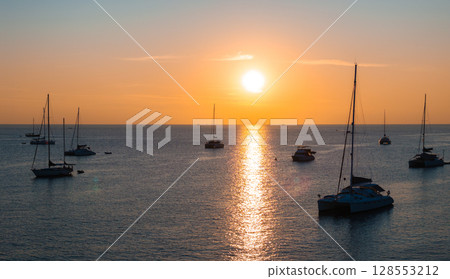 Serene aerial view of Naiharn Beach in Phuket, Thailand, at sunset. The calm sea reflects the golden light, with sailboats and yachts anchored nearby. Serene aerial view of Naiharn Beach in Phuket, Thailand, at sunset. The calm sea reflects the golden light, with sailboats and yachts anchored nearby. 128553212