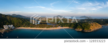 Aerial view of Naiharn Beach in Phuket, Thailand, featuring a crescent shoreline, lush green hills, a lagoon, white buildings, and distant islands. 128553223