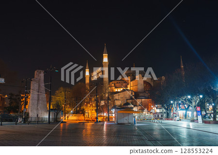 Hagia Sophia's domes and minarets glow at night in Istanbul, Turkey. A lighted Arabic message, trees, a paved square, and a stone column are visible. Hagia Sophia's domes and minarets glow at night in Istanbul, Turkey. A lighted Arabic message, trees, a paved square, and a stone column are visible. 128553224