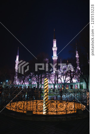 The Sultan Ahmed Mosque, with domes and six minarets, glows at night in Istanbul. The Serpent Column stands in the foreground, framed by trees. 128553228