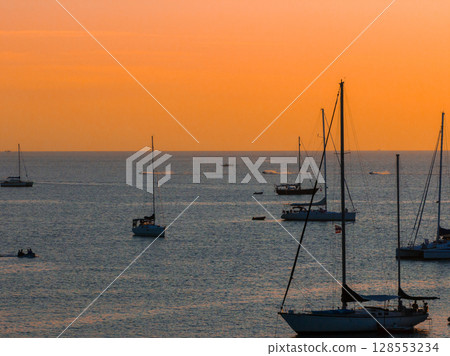 Sunset at Naiharn Beach in Phuket, Thailand, with orange skies reflecting on calm waters. Sailboats and vessels float peacefully in the tranquil scene. Sunset at Naiharn Beach in Phuket, Thailand, with orange skies reflecting on calm waters. Sailboats and vessels float peacefully in the tranquil scene. 128553234