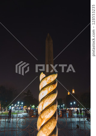 Night view of the Serpent Column and Obelisk of Theodosius in Sultanahmet Square, Istanbul, with warm lighting, minaret silhouettes, and lively activity. 128553235