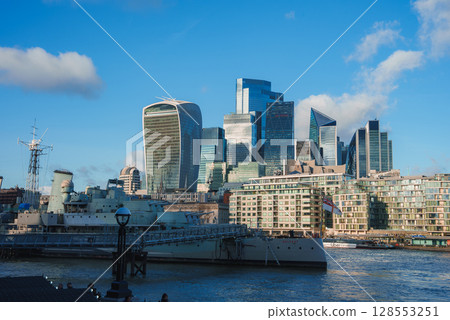 The iconic Walkie Talkie building and HMS Belfast on the River Thames, with modern skyscrapers like the Leadenhall Building under a clear blue sky. 128553251