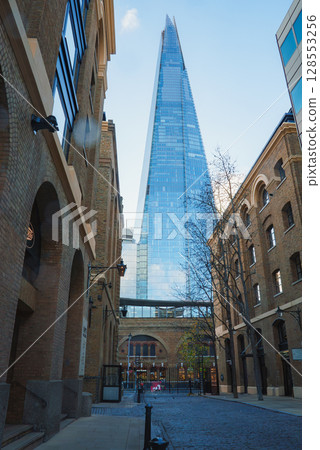 The Shard rises above a narrow cobblestone street in London, contrasting with historic brick buildings and bare trees under a clear winter sky. 128553256