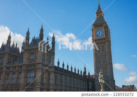 The iconic Big Ben and Gothic architecture of the Houses of Parliament stand prominently under a clear blue sky with scattered clouds in London, England. 128553272