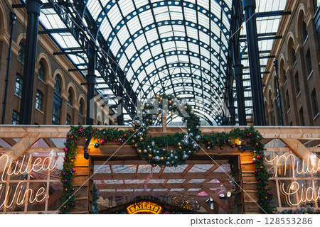 Hay's Galleria in London features an arched glass roof and festive decor. A wooden stall with a wreath and neon signs offers mulled wine, creating a warm atmosphere. Hay's Galleria in London features an arched glass roof and festive decor. A wooden stall with a wreath and neon signs offers mulled wine, creating a warm atmosphere. 128553286