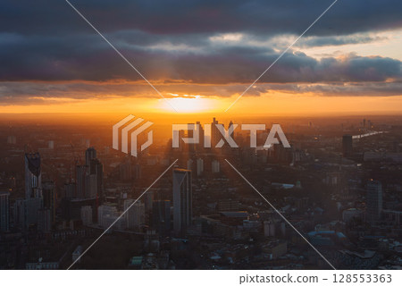 The image shows a sunset over London with Canary Wharf skyscrapers, a golden glow on the horizon, dark clouds, and the River Thames winding through the city. The image shows a sunset over London with Canary Wharf skyscrapers, a golden glow on the horizon, dark clouds, and the River Thames winding through the city. 128553363