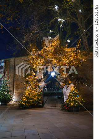 A festive archway in London adorned with twinkling lights, golden ornaments, and greenery. A lamppost and lit Christmas trees enhance the holiday scene. 128553365