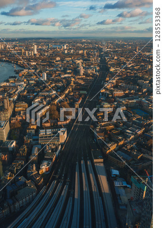 Aerial view of London during Christmas, featuring railway tracks, the River Thames, and city landmarks under a partly cloudy, golden sky. Aerial view of London during Christmas, featuring railway tracks, the River Thames, and city landmarks under a partly cloudy, golden sky. 128553368