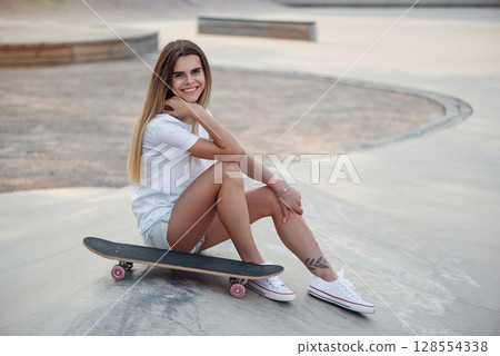 Attractive girl with a tattoo in a white t-shirt sitting on a ramp in skate park with a skateboard. Attractive girl with a tattoo in a white t-shirt sitting on a ramp in skate park with a skateboard. 128554338