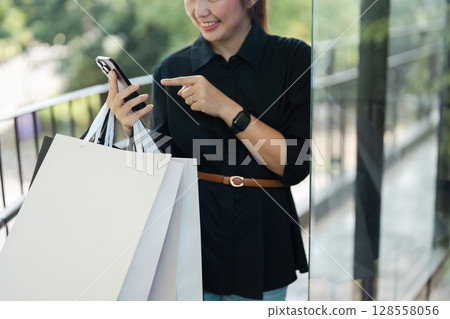 Woman Shopping with Bags and Smartphone, Smiling and Pointing at Screen, Modern Retail Experience Woman Shopping with Bags and Smartphone, Smiling and Pointing at Screen, Modern Retail Experience 128558056