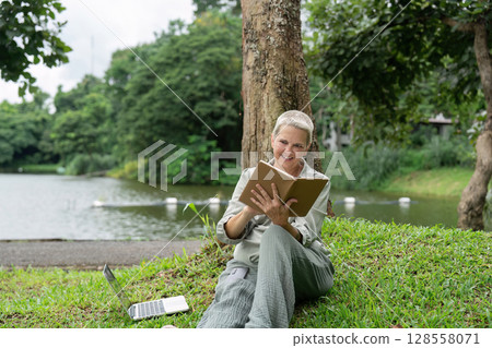 Senior Woman Enjoying a Digital Tablet Outdoors by a Lake in a Park Setting with Greenery and Trees Senior Woman Enjoying a Digital Tablet Outdoors by a Lake in a Park Setting with Greenery and Trees 128558071