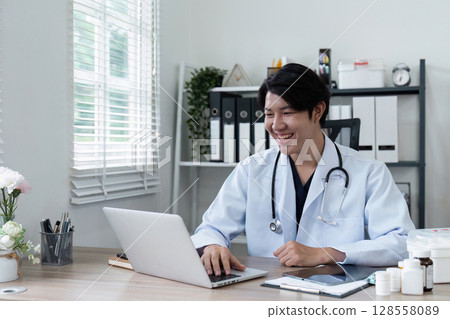Smiling Doctor Working on Laptop in Modern Office with Medical Equipment and Documents Smiling Doctor Working on Laptop in Modern Office with Medical Equipment and Documents 128558089