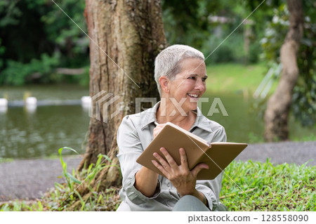 Senior Woman Enjoying a Book Outdoors in a Park by a Lake on a Sunny Day 128558090