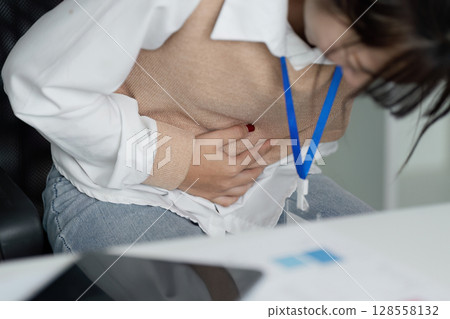 Office Worker Feeling Sick and Unwell at Desk, Holding Stomach in Pain, Experiencing Discomfort During Work Hours 128558132
