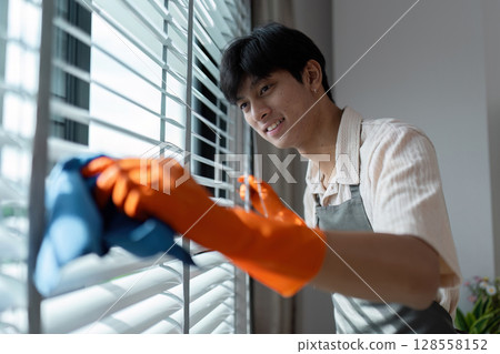 Young Man Cleaning Window Blinds with Orange Gloves and Blue Cloth in a Modern Home Interior 128558152