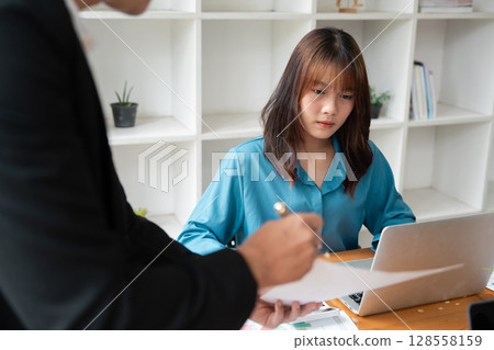 Young Professional Woman Working on Laptop in Modern Office with Colleague Reviewing Documents 128558159